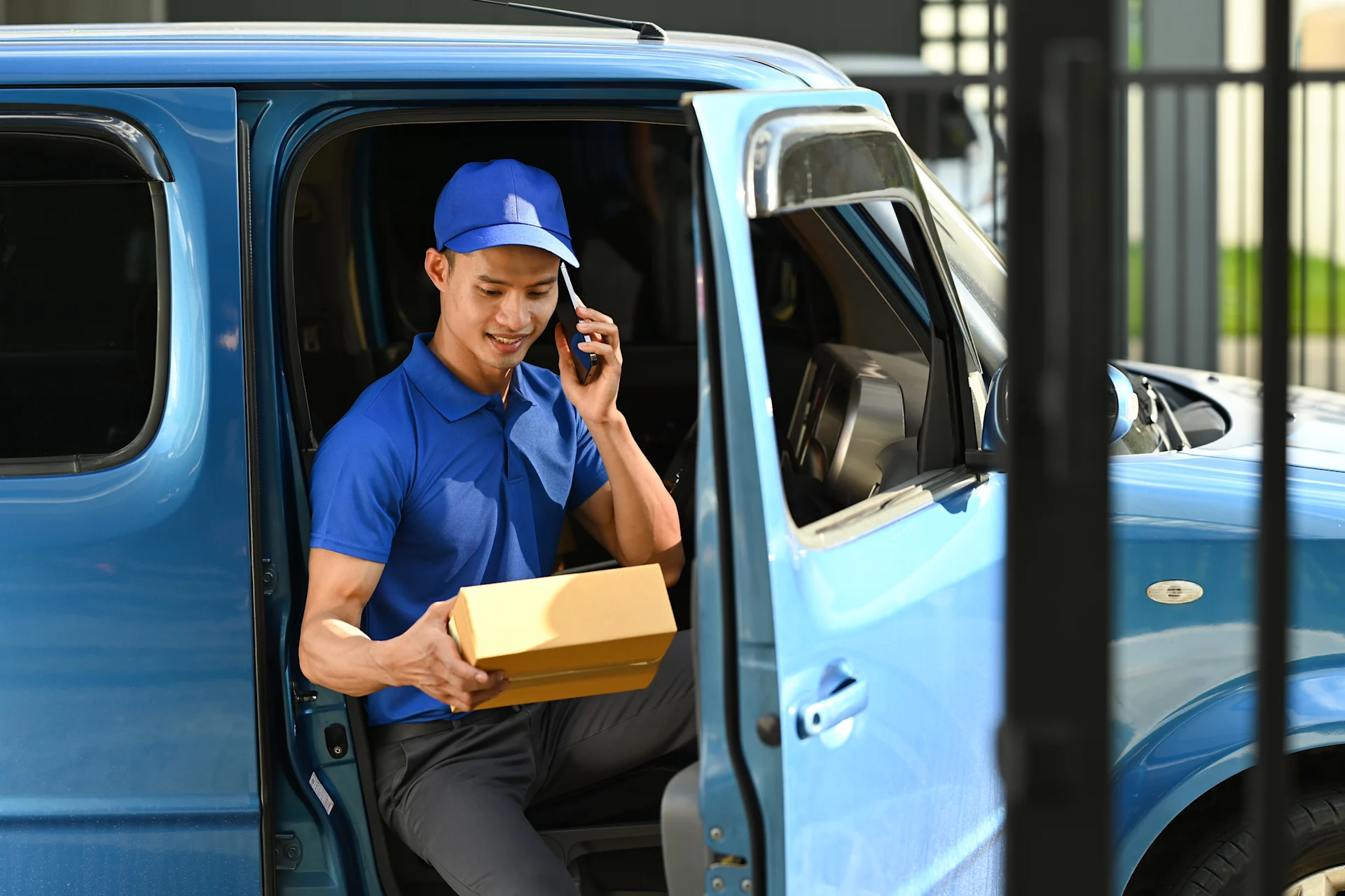 Delivery driver in blue outfit getting out of blue van with box in one hand and phone in the other